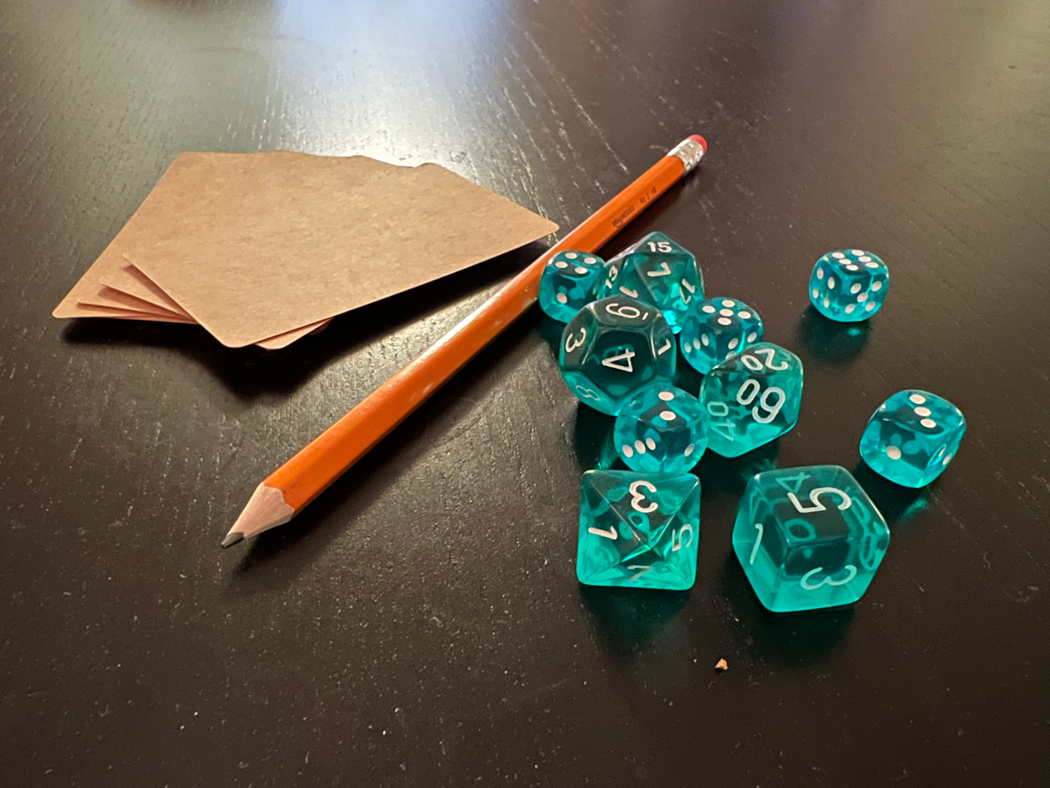 A stack of cardboard cards, a number 2 pencil, and a handful of semi-translucent cyan blue dice sit on top of a dark brown table to signify common household objects you can use to prototype your gameplay.