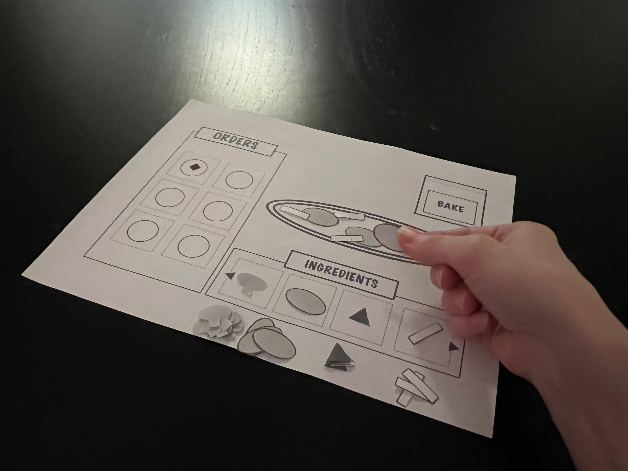 A lightskinned hand holds a small paper cutout of a piece of pepperoni over a piece of paper with a UI prototype of a pizza building system. The piece of paper is on a dark brown table.